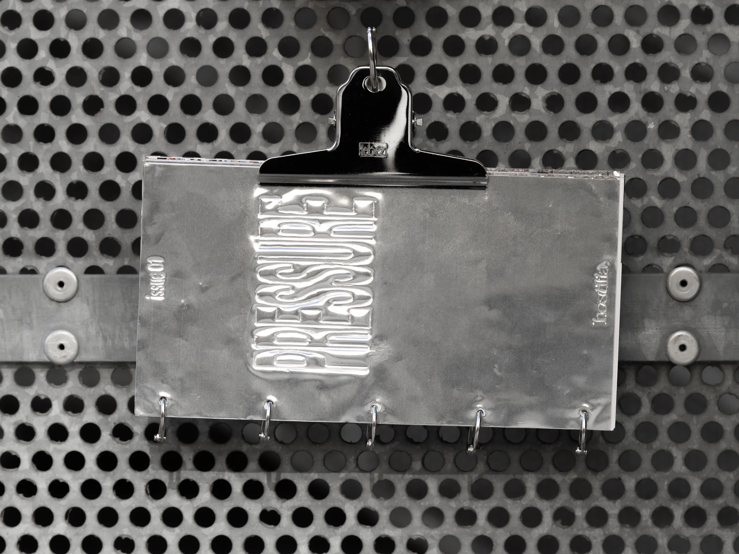 The aluminium cover of Hostilia, embossed with the word Pressure, held by a clip on a perforated metal sheet