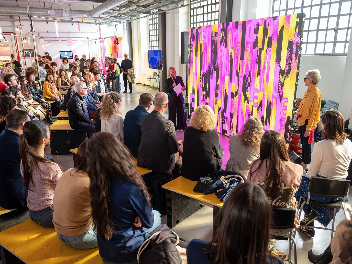 The audience of a talk, seated beside a large yellow and magenta panel of the identity