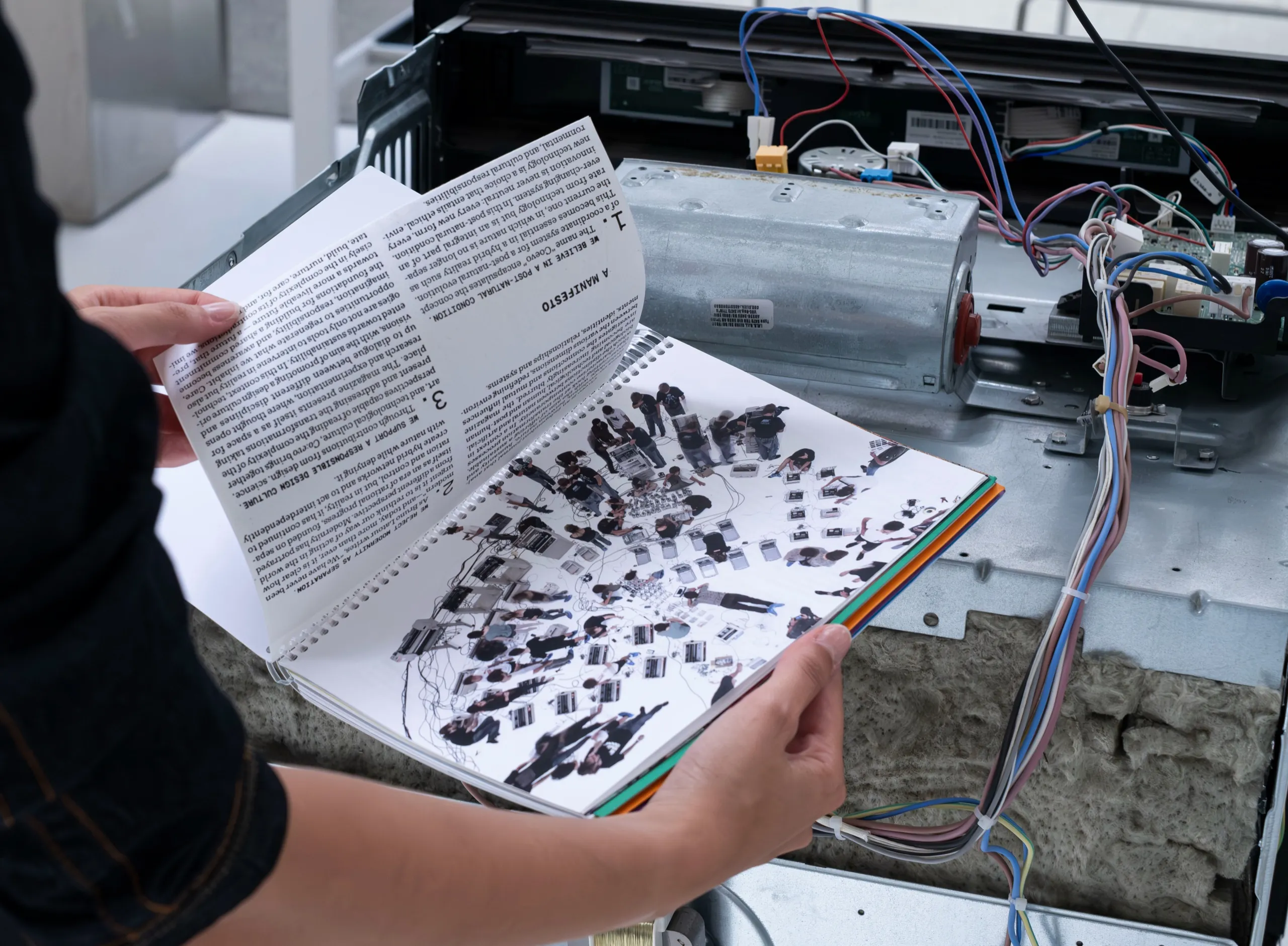 Hands opening the fold-out manifesto of co:evo in front of an open machine with exposed wiring