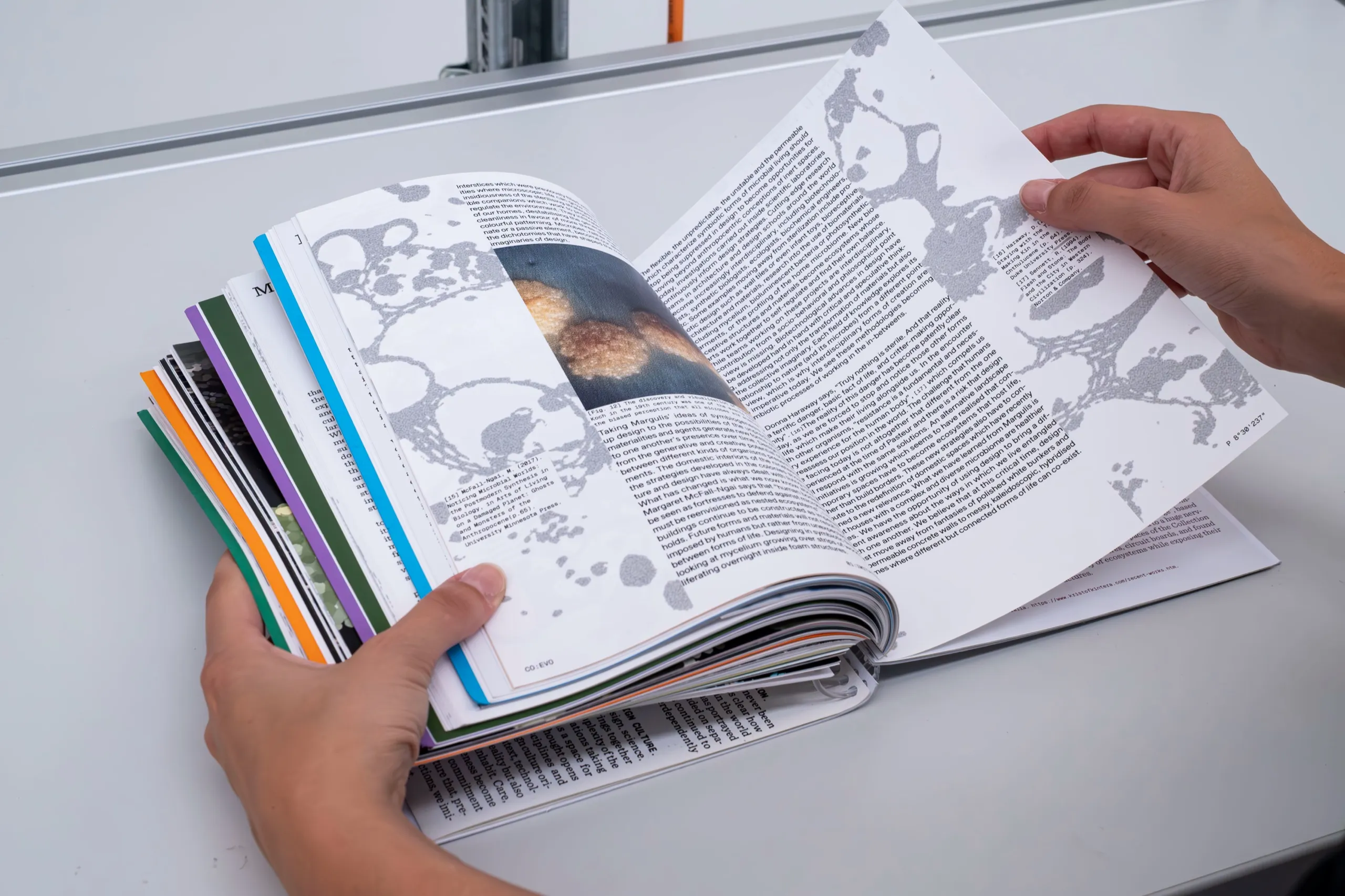 Hands leafing through co:evo, open on a text page with grey organic shapes, on a white work surface